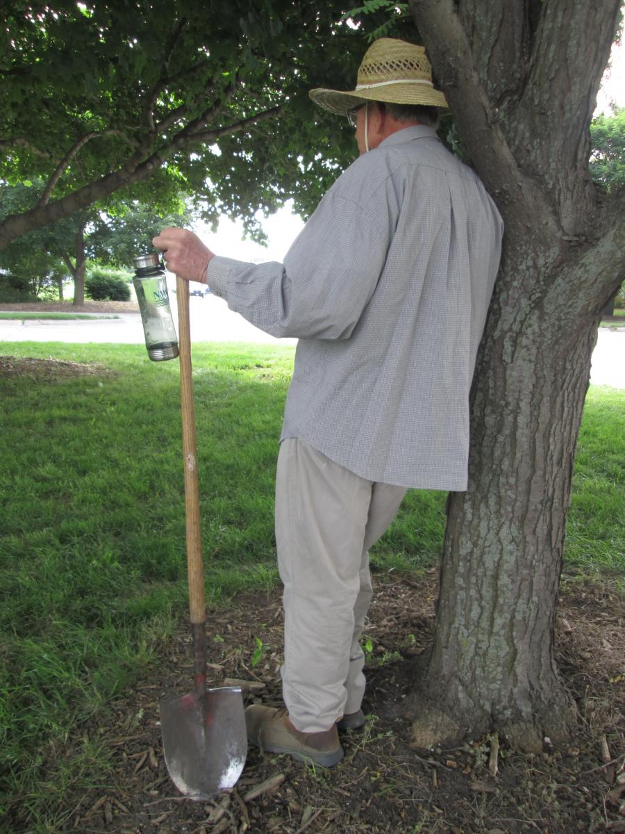 A man wearing light-colored clothing standing with a water bottle in the shade of a tree