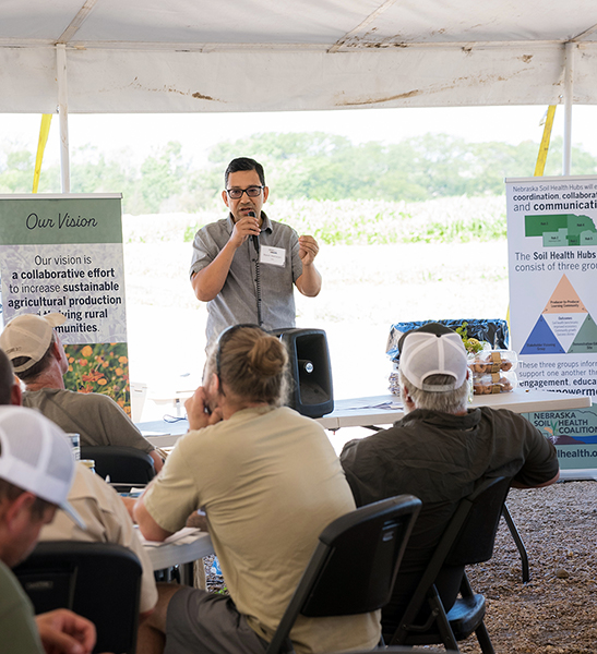 Bijesh Marharjan holds a microphone and addresses attendees seated under a tent at a Nebraska Soil Health Coalition event, with educational posters and crop fields visible in the background.