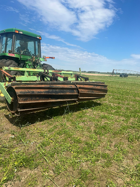 Green tractor with rusted farm equipment on a grassy field under a bright blue sky.
