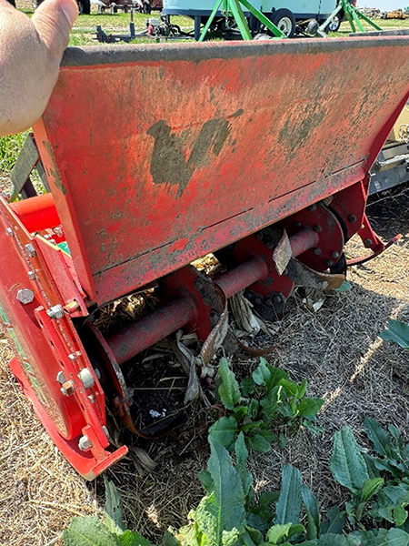 Rusty red agricultural equipment in a field, surrounded by weeds and dry grass.