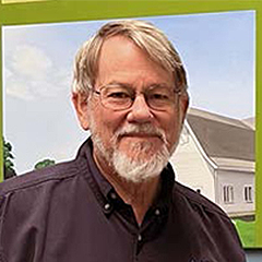 Headshot of Corey Brubaker in dark-colored shirt standing in front of painting on wall and smiling at camera