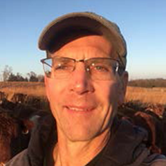 Headshot of Doug Garrison in brown jacket and hat amidst cattle in field during fall