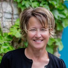 Headshot of Angela Knuth in black top with green foliage and stone wall in the backgorund