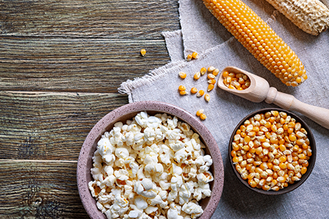 Bowl of popped popcorn beside bowls of yellow popcorn kernels, whole corn cobs and a wooden scoop on a rustic wood surface.