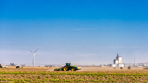 John Deere green and yellow tractor kicks up dust through a corn residue field under bright blue sky, with grain silos and center pivot in the distance