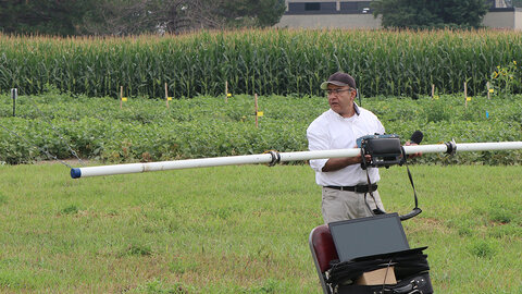 A University of Nebraska–Lincoln researcher stands in a crop field holding a large sensor instrument connected to a laptop, preparing to collect geophysical data to assess soil and water conditions near an irrigation research site.