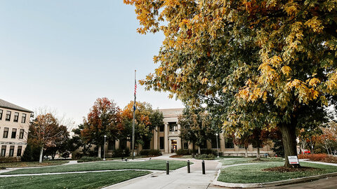 Fall trees with orange and yellow leaves frame a campus walkway leading to a stately university building under a clear sky.