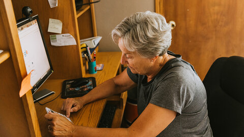Woman seated at a desk using a calculator beside a computer, reviewing paperwork in a home office.
