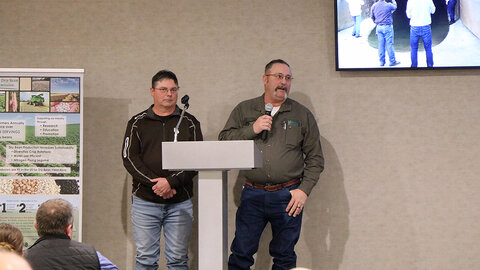 Two men stand at a podium during a conference presentation, one speaking into a handheld microphone, with a slide and dry bean display behind them.