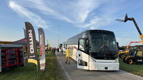 Bus parked on a paved road at an outdoor event with flags and machinery nearby.