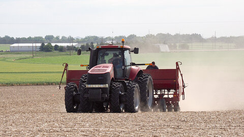 Red tractor pulling farming equipment through a dusty field with green farmland in the background.