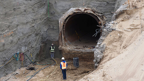 Tunnel excavation site with two workers wearing safety gear, surrounded by dirt and equipment.