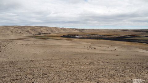 Expansive barren landscape with sandy hills, sparse vegetation, and cloudy sky.