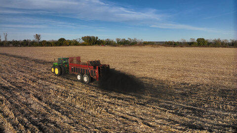 A tractor pulls a manure spreader across a harvested field, applying biochar over crop residue under a blue sky.