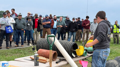 A person demonstrating tools to a large group outdoors under a cloudy sky.
