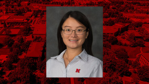 Head-and-shoulders portrait of Yeyin Shi wearing glasses and a striped shirt with a red Nebraska “N” logo, set against a red aerial campus background.