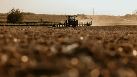 Closeup of soil in field with planter kicking up dust in the background