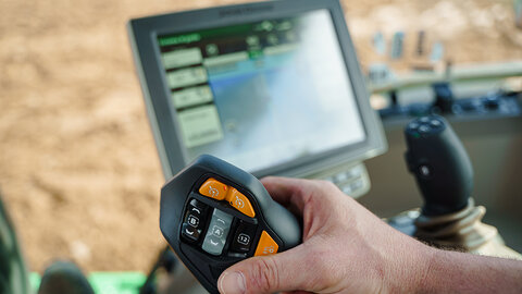 A farmer’s hand holds a precision agriculture control handle inside a tractor cab, with a touchscreen display showing field data and GPS mapping in the background.
