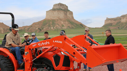 A group of people gathered around an orange tractor in a rural landscape with rocky hills.