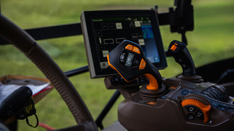 Close up of a tractor cab shows a joystick with orange accents, an auxiliary stick, and a touchscreen console running guidance and implement tools in soft daylight.