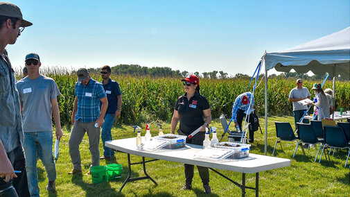 Woman stands at table in front of field day attendees near field