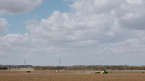 Wide view of Nebraska farmland under a partly cloudy sky, with a tractor and irrigation system working in a freshly tilled field, and wind turbines standing in the distance beyond a line of trees.          Ask ChatGPT