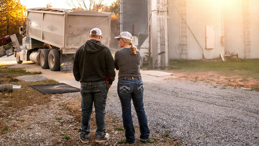 A man and woman in ball caps stand together near grain bins and a semi-truck unloading corn on a farm at sunset.