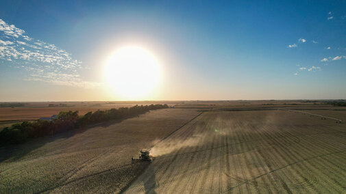 A combine harvests crops in a wide Nebraska field at sunset, leaving a trail of dust under a clear sky.