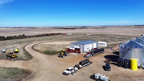 Aerial view of a farmstead with grain bins, equipment sheds, semitrucks, tractors, and pickup trucks parked around the yard, surrounded by expansive open fields under a clear blue sky.