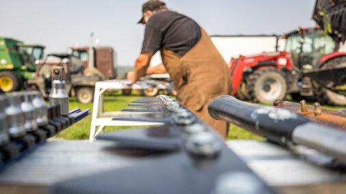 Close-up of a workbench with tools and machine parts laid out in the foreground, while a farmer in brown coveralls performs maintenance on equipment in a field with tractors and harvest machinery in the background