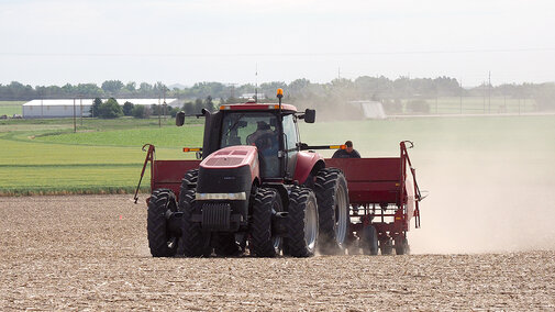 Red tractor pulling farming equipment through a dusty field with green farmland in the background.