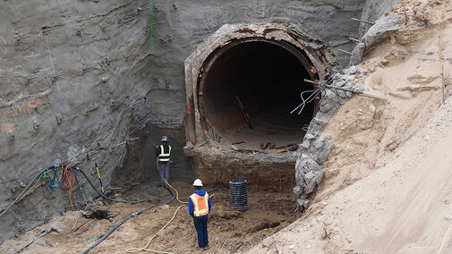 Tunnel excavation site with two workers wearing safety gear, surrounded by dirt and equipment.