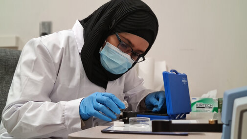 Scientist in lab coat and gloves working with lab equipment at a table.