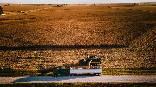 Grain truck drives on country road alongside combine that pours corn grain into bin as it harvests corn field 