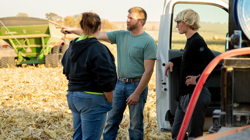 Group talks near truck on harvested field, green equipment in background, sunny day.