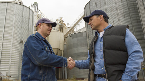 Two farmers standing in front of grain bins shaking hands