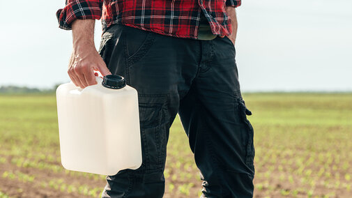 Person holding an unmarked pesticide container in an early-season cornfield, illustrating herbicide planning and compliance requirements.