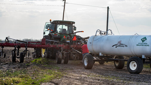 Tractor pulling an anhydrous ammonia tank and applicator toolbar in a field, preparing nitrogen fertilizer for crop production.