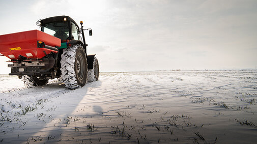 Tractor with a red spreader drives across a snow-covered field, with tire tracks visible and sparse green plants poking through the snow.
