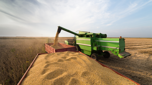 A combine unloads freshly harvested soybean into a grain cart in the middle of a field under a partly cloudy sky. The harvested field stretches into the distance, and the grain cart is nearly full of golden soybean.