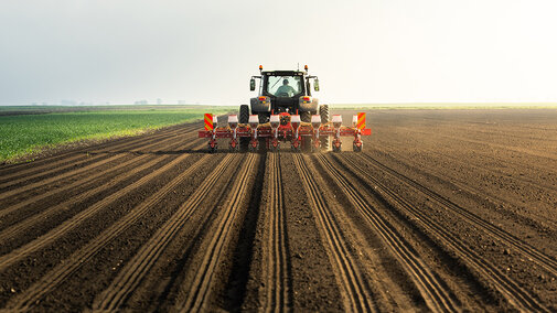 Tractor planting seeds in a vast, freshly plowed field under a bright sky.