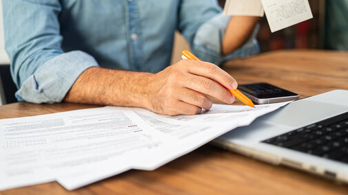 Person reviewing paperwork and receipts with pen at desk beside laptop.