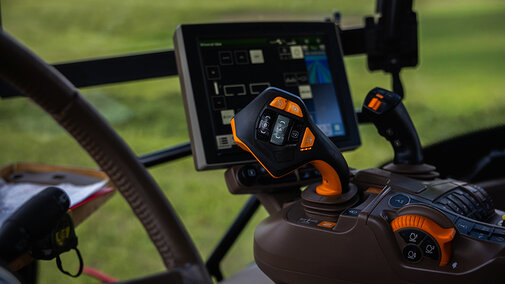 Close up of a tractor cab shows a joystick with orange accents, an auxiliary stick, and a touchscreen console running guidance and implement tools in soft daylight.