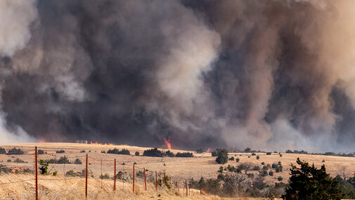 Wildfire burning across dry pastureland with flames and thick smoke rising behind a fence line in a rural landscape.