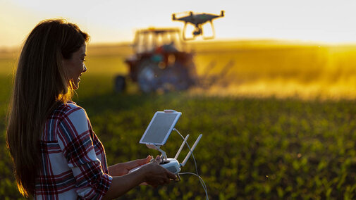 farmer operates drone with handheld console as it flies into soybean field while tractor drives in background