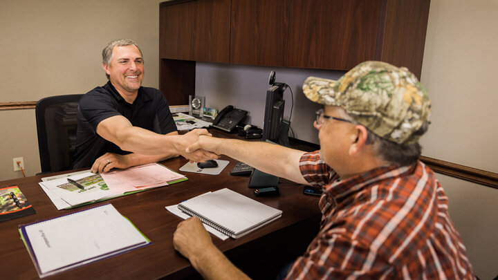 man in casual apparel shaking hands with man in professional apparel over desk