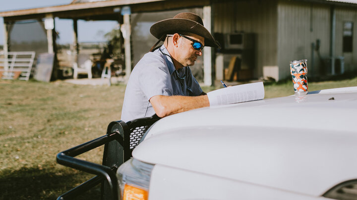 A farmer reviews paperwork on the hood of a pickup truck outside a rural building, illustrating on-farm decision-making or succession planning activities.
