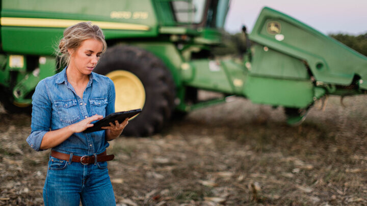 Woman in denim using a tablet in a harvested cornfield, standing beside a green combine — illustrating technology use in modern farm management.