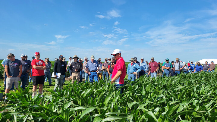 Group of farmers and ag professionals gathered in a cornfield during the 2025 weed management field day, listening to a speaker in a red Nebraska shirt under a bright blue sky.