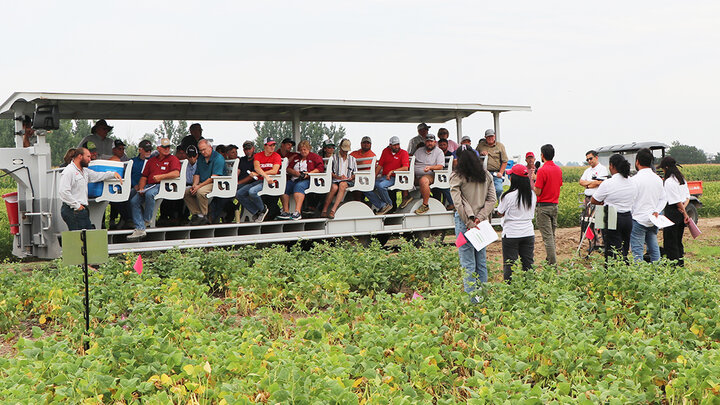 A group of people ride on a covered field tour trailer through a research plot of green bean plants, listening to a presenter at the front. Additional attendees stand nearby holding handouts, with crop rows and other research plots in the background.